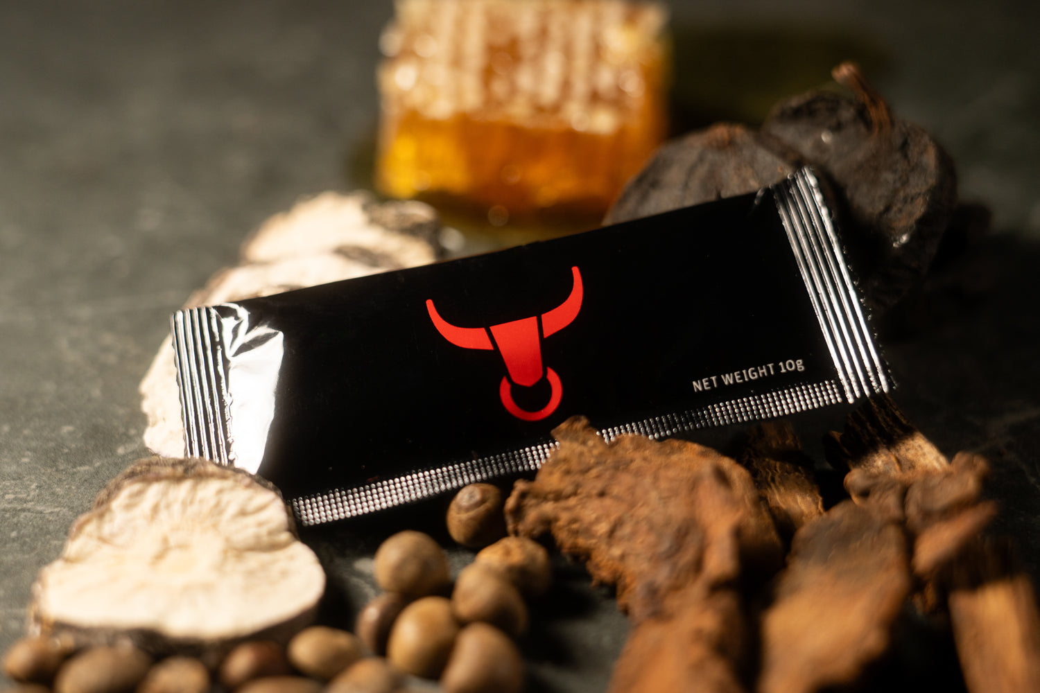 A black supplement powder sachet with a red bull logo and 'NET WEIGHT 10g' printed on it, styled on a dark surface with coffee beans, dried herbs, and honey in the background.